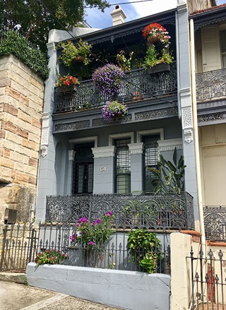 Front view of grey double story terrace house with coloured pot plants hanging from upstairs verandah with a dark grey lacy fence.