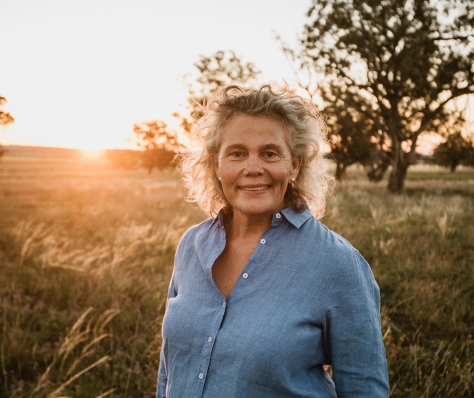 Biodiversity Conservation Trust member Fiona Simson, wearing a blue shirt and standing in a field with trees and the sun behind her