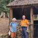 An elderly couple stand in front of a small stone hut with a thatched roof and log frame in a clearing in a forest