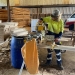 Someone in fluorescent work gear and a cap that says "crusty demons" stands cutting wood; there are large stacks of slats of different shapes behind him in a large corrugated iron shed