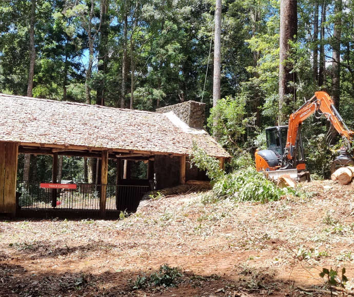 A wooden hut with a stone chimney and shingled roof in a cleared area by a forest, with an orange construction vehicle picking up logs in the foreground