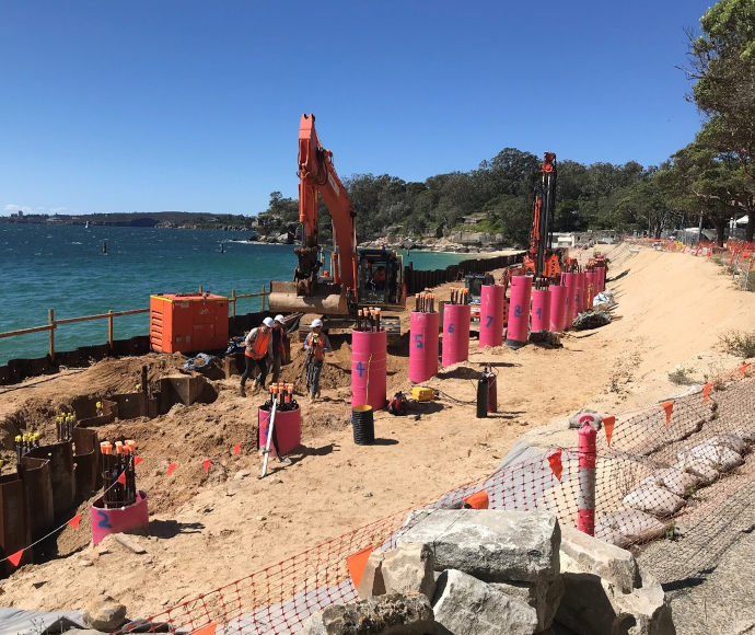 Construction machines and three workmen in helmets and fluorescent vests in a trench on the beach at Nielsen Park, with a blue ocean, blue sky and thickly treed point in the background