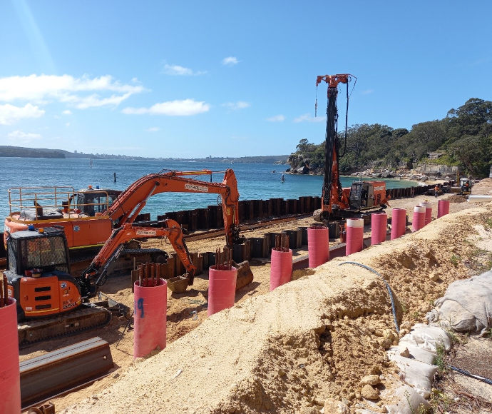 Construction machines and equipment in a trench in the sand on the beach at Nielsen Park, with a blue ocean, blue sky and thickly treed point in the background