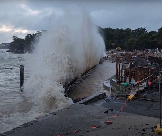 A grey sea under low-hanging grey clouds; a jet of sea spray rising up on impact with a low seawall around some construction works