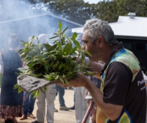 Man blowing smoke from flowering gum leaves, held in a carved wooden board filled with ash