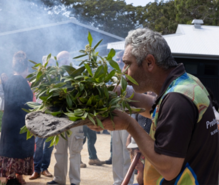Man blowing smoke from flowering gum leaves, held in a carved wooden board filled with ash