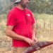 A man in a red t-shirt holds a finely carved, wooden Aboriginal artefact with finely engraved patternings.