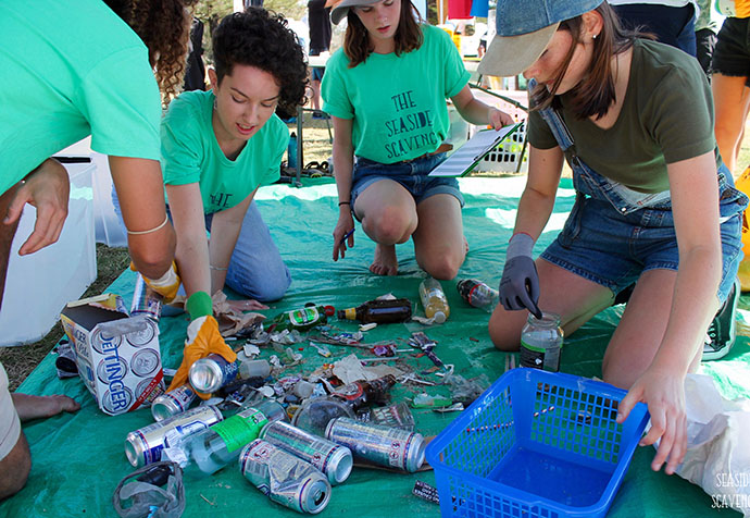 Four Sea Scavenge volunteers kneeling on green tarpaulin in a circle sorting rubbish. Aluminium cans and other rubbish in centre and blue plastic tub in foreground.