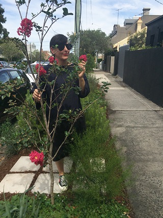 Woman in sunglasses standing in her verge garden next to footpath holding flower of one of her plants.