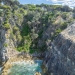 Aerial view of a small clear-watered inlet protected on all sides by grey cliffs, rocky outcrops and thick green scrub