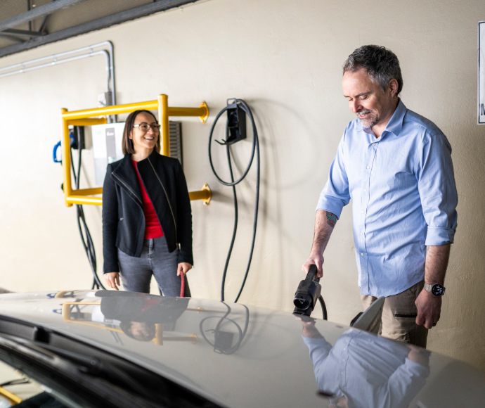 Smiling man charging an electric vehicle in an apartment building that has been retrofitted with a charging station, next to him is a woman with a whippet or greyhound on lead but just out of shot