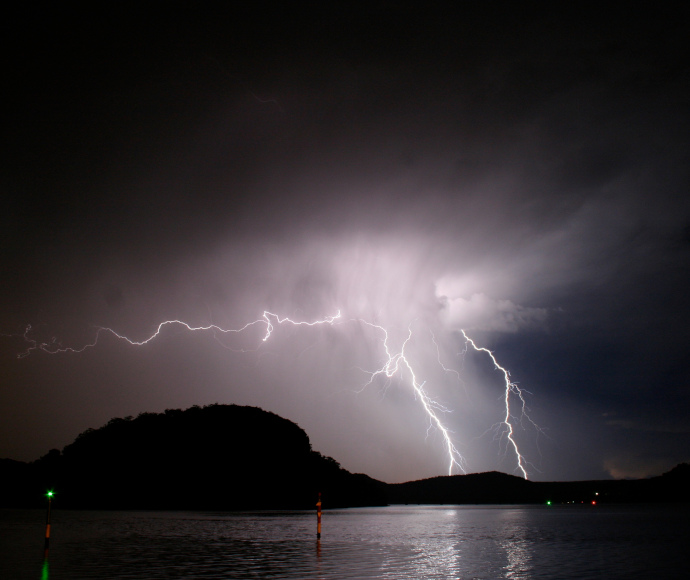 A night-time scene of lightning striking thickly treed hills facing the dark waters of a bay; the water, sky and clouds are lit by white lightning