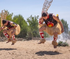 Two young dancers in ceremonial dress photographed at a moment when they are both about a foot off the ground, their own noontime shadows directly beneath, with ceremonial smoke in the background