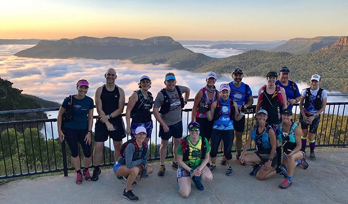 A group of runners stand on a lookout platform with the view of mountains and valleys in the background, within Blue Mountains National Park. 