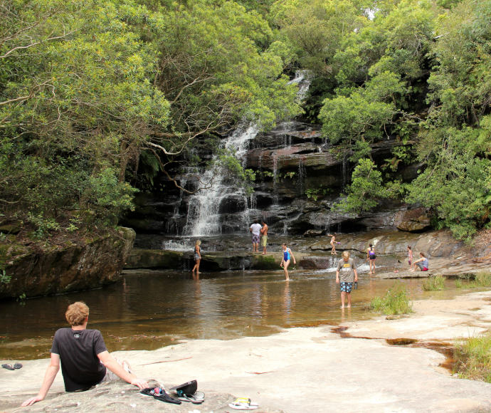 Person relaxing on rocks watching others in rockpool below waterfall