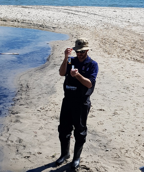 Person taking water sample from drain flowing onto a beach