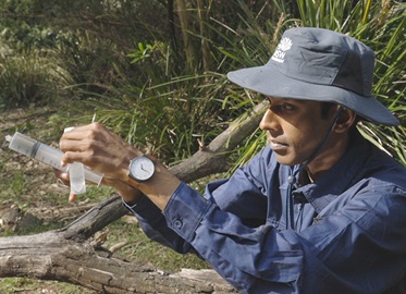 Scientist in blue shirt and grey hat holds a syringe used for water quality testing at Wapengo Creek, Wapengo Lagoon Catchment 