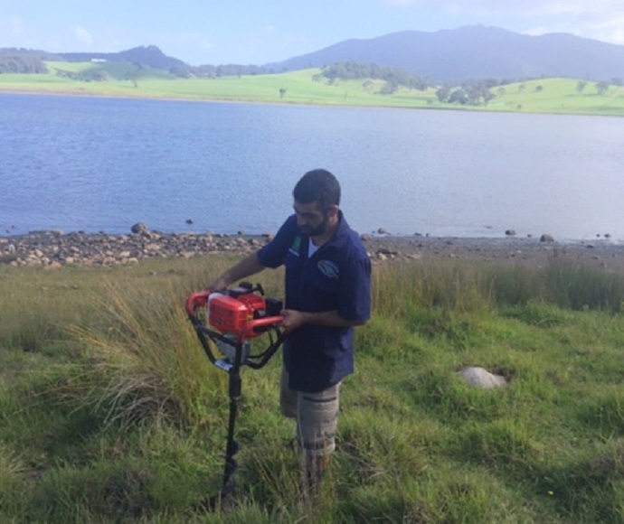 Person using an auger to dig a hole to plant a tree, with Tilba Tilba Lake in the background