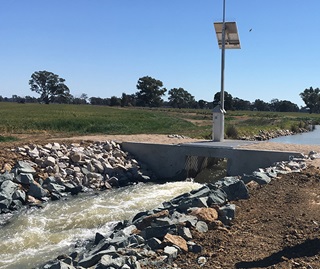 Release of water to Tuppal Creek from the Murray Irrigation Limited escape. Concrete platform across middle of creek with stones on creek banks and rushing water in foreground. 