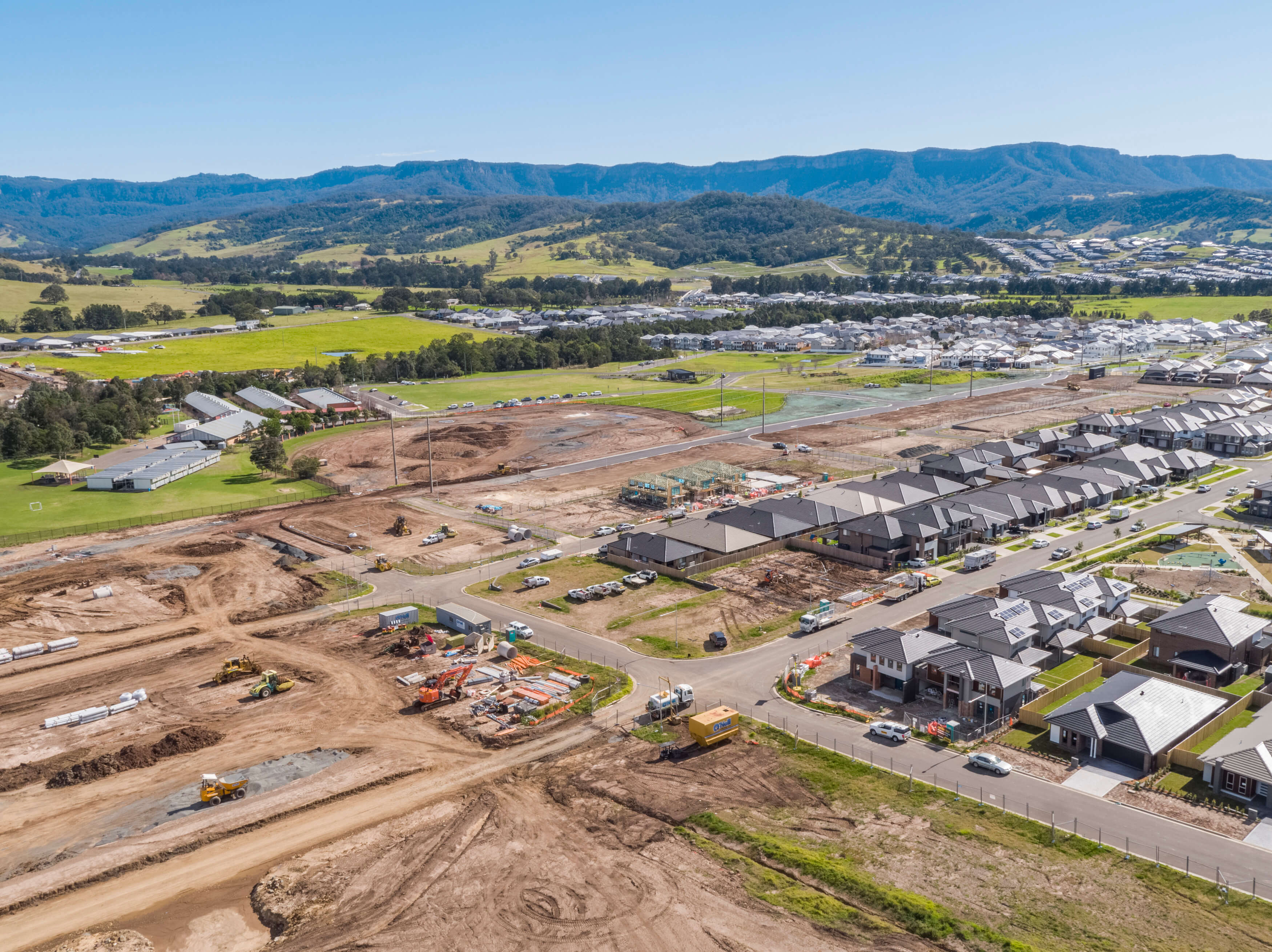 View shows subdivisions and buildings under construction and areas of grassland, trees and hills in distance