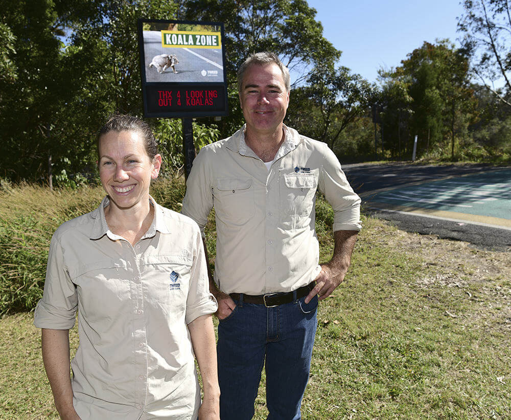 Scott Hetherington and Marama Hopkins, of the Tweed Shire Council, standing at the side of a road with a large sign that reads - Koala Zone.