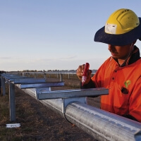 Worker in an orange uniform and yellow safety helmet marking a metal structure in an open field, with a clear sky and colleagues in the background.