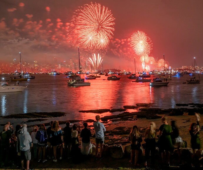 A vibrant fireworks display over Sydney Harbour, with multiple boats and a crowd of spectators gathered on the shore at night.