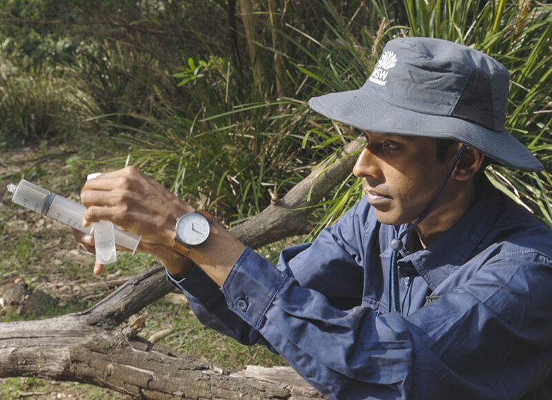 Scientist in blue shirt and grey hat holds a syringe used for water quality testing at Wapengo Creek, Wapengo Lagoon Catchment