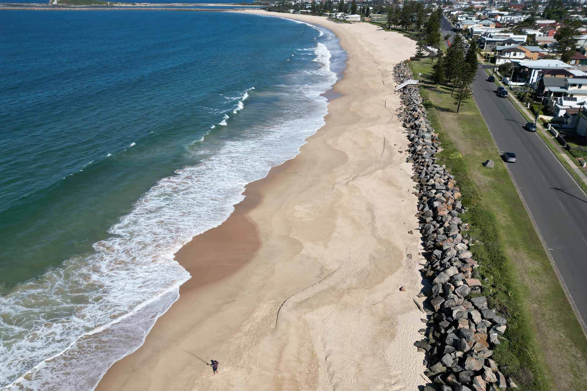 Aerial view of a sandy beach with waves lapping the shore next to a tree-lined street, fisherman in foreground