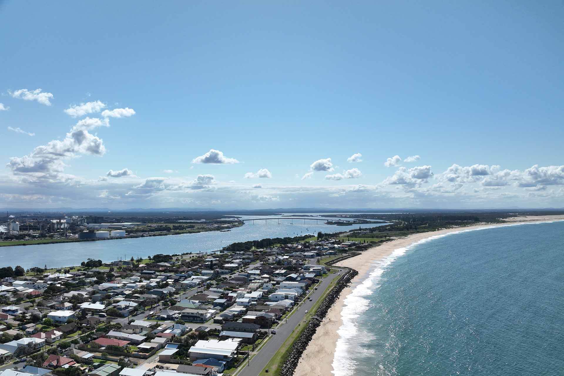Aerial view of a large dredging ship near a coastline, expelling water with a populated area in the background.