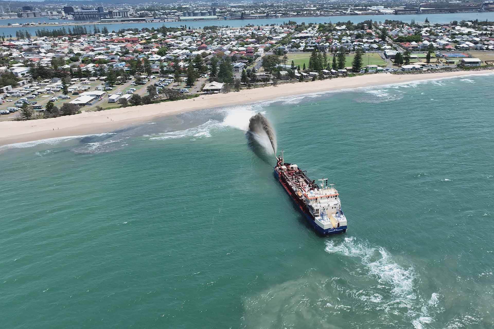 Aerial view of a large dredging ship near a coastline, expelling sand with a populated area in the background