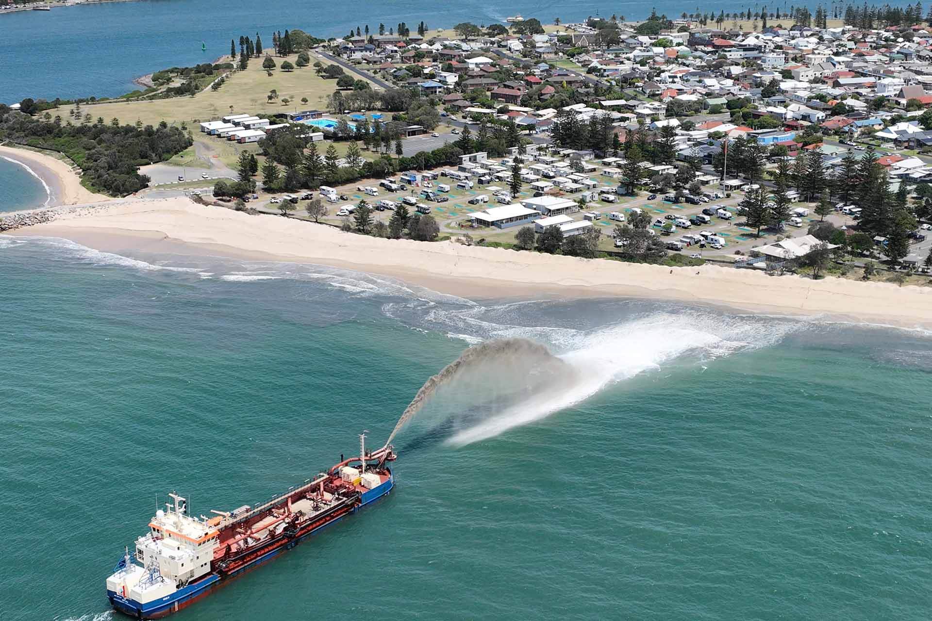 Aerial view of a large ship discharging sand near a coastal area with buildings and vegetation