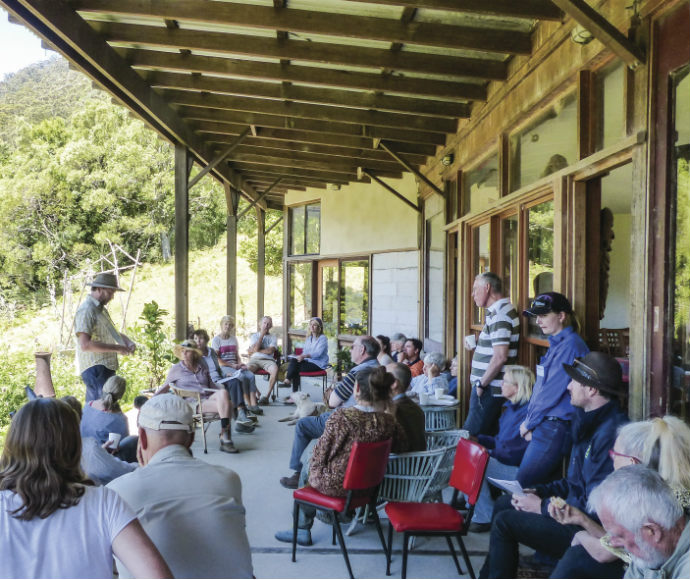 a group of people gathered on a covered porch of a rustic building, listening to an individual who is standing and addressing the group. The setting appears to be in a rural or natural area with greenery visible in the background. There are various seating arrangements, including folding chairs and benches. The architecture features wooden beams and supports, with the exterior wall of the building showing aged wood siding.