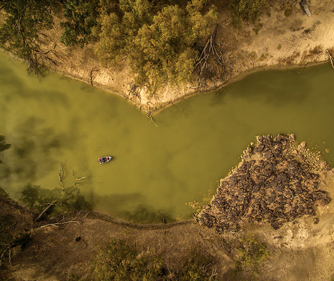 An aerial view of the Darling River at the Darling River campground in Toorale National Park. Trees line both sides of the river, and a small boat is visible on the water. The photo has a greenish-yellow tint, giving it a unique and vibrant appearance.