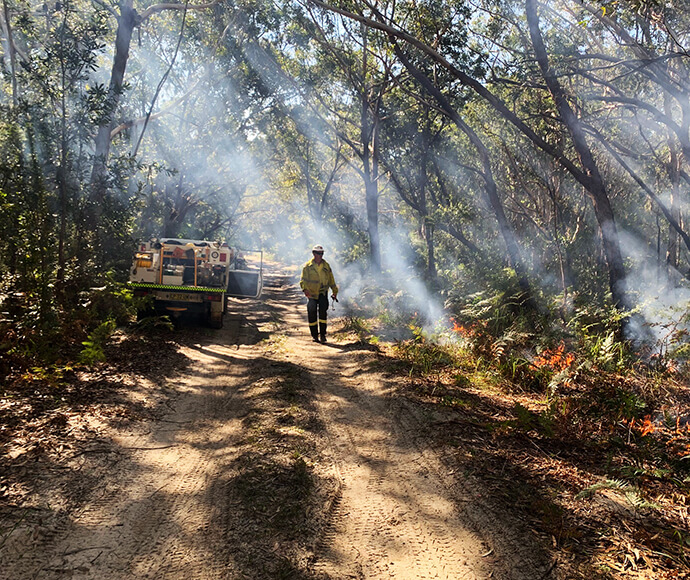 A firefighter in protective clothing walking away from the camera along a dirt track during a hazard reduction burn at Treachery Beach. Low flames can be seen in the vegetation to the right and there is smoke through the trees.