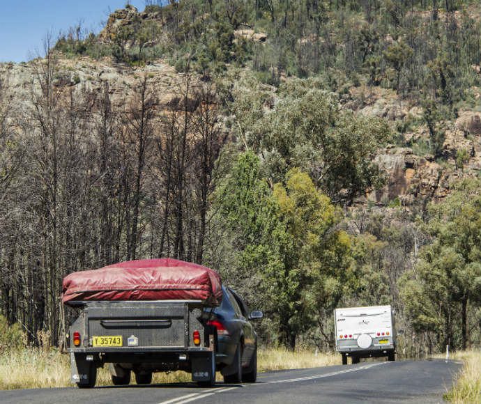 A black pickup truck with a red cover over its cargo bed drives on a rural or mountainous road. Ahead, a white camper trailer is being towed by another vehicle. The landscape features grassy areas, dense trees, and rocky hills under a clear sky.