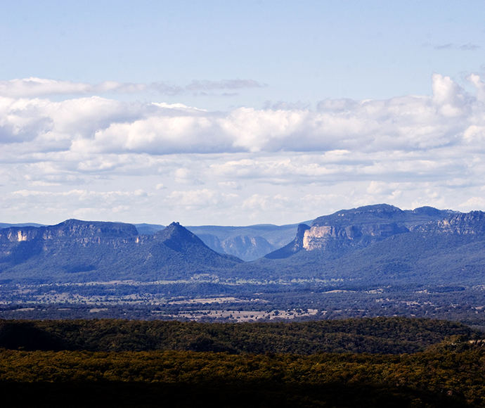 A breathtaking view from Pearsons Lookout in Gardens of Stone National Park. The foreground features a railing and a rocky platform, while the background showcases an expansive landscape of rugged cliffs, deep valleys, and dense forests under a partly cloudy sky. The dramatic rock formations and vibrant greenery create a stunning and natural scene.