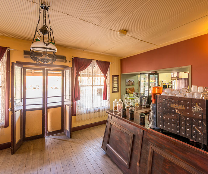 The interior of a historic general store and café at Hill End Historic Site, featuring rustic wooden shelves stocked with various goods, including canned items, jars, and vintage-style products. A wooden counter with a simple cash register is in the foreground, and a chalkboard menu is displayed on the wall. The space is warmly lit, creating a cozy, nostalgic atmosphere. Wooden beams and antique décor enhance the vintage charm, giving visitors a glimpse into the store’s historic ambiance and old-world charm