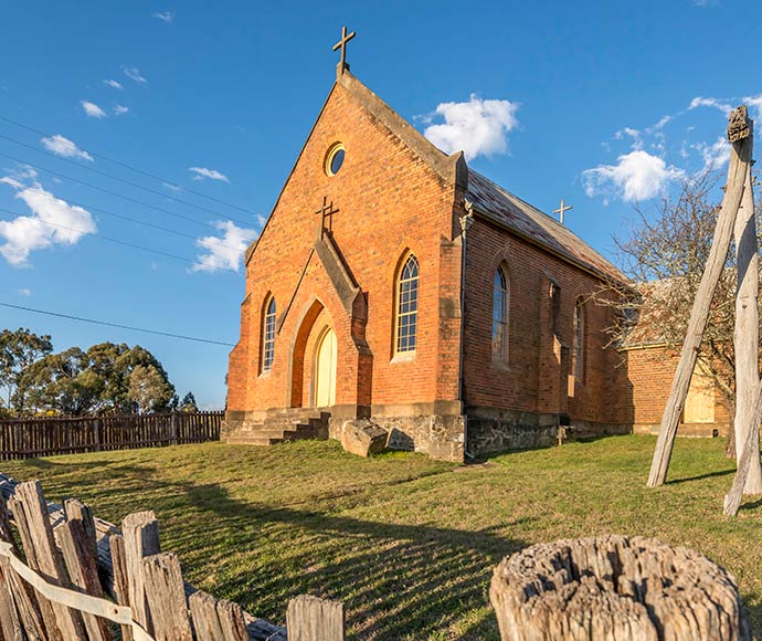 The exterior of an old Catholic church on Denison Street at Hill End Historic Site, featuring rustic red brick walls and a simple, pitched roof. The small church has arched windows with white trim, and a modest entrance with a white-framed door. Surrounded by a grassy yard with a few trees and shrubs, the church is set against a backdrop of greenery and a clear sky, evoking a sense of tranquility and historical charm. The scene captures the quiet, preserved character of this historic place of worship.