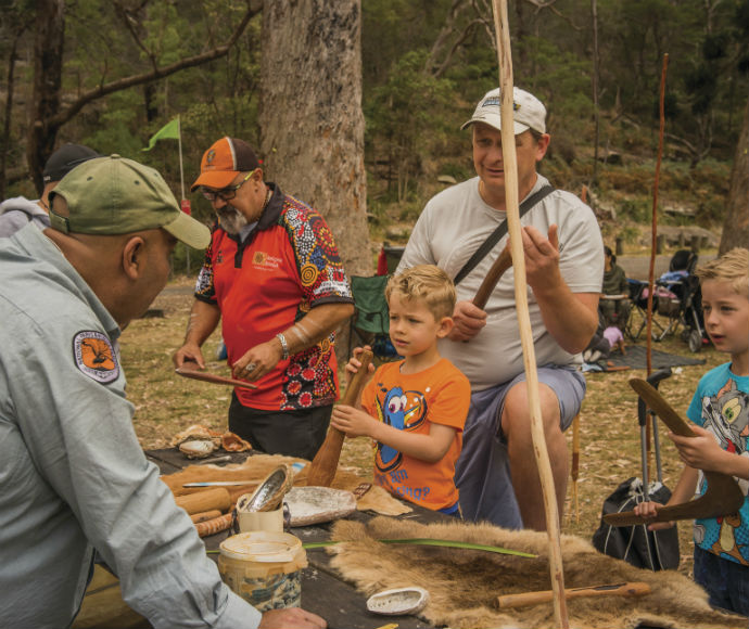 A small group participates in an Aboriginal cultural heritage workshop at Royal National Park. Participants gather around an instructor who shares traditional knowledge and practices, surrounded by natural bushland. The workshop aims to foster understanding and respect for Aboriginal culture and connection to Country.