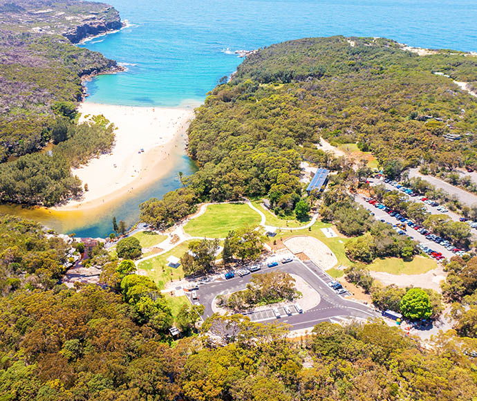 Aerial view of the Wattamolla picnic area in Royal National Park. The image highlights a green lawn area with multiple picnic tables and shelters, surrounded by dense eucalyptus forest. There is a parking lot filled with cars near the bottom of the image. A lagoon is visible, connected to the ocean by a narrow strip of beach. The scene is serene and well-maintained, showcasing a popular recreational spot with both water and forest elements.