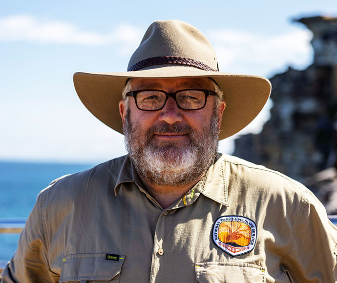Deon van Rensburg stands in the foreground wearing his National Parks and Wildlife Service uniform, against a stunning ocean backdrop.