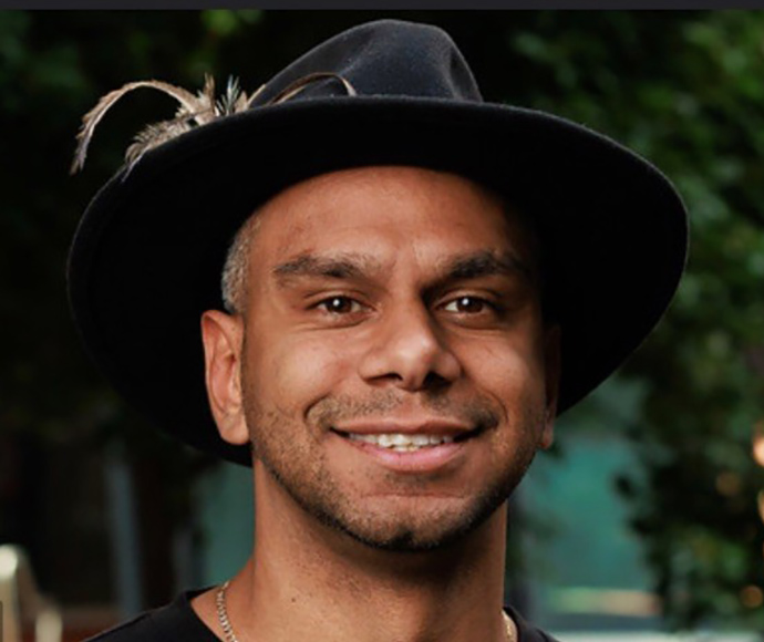 Jonathon Captain-Webb smiles proudly while wearing a black felt hat with a feather. He appears to be in a natural landscape although the background is blurred.