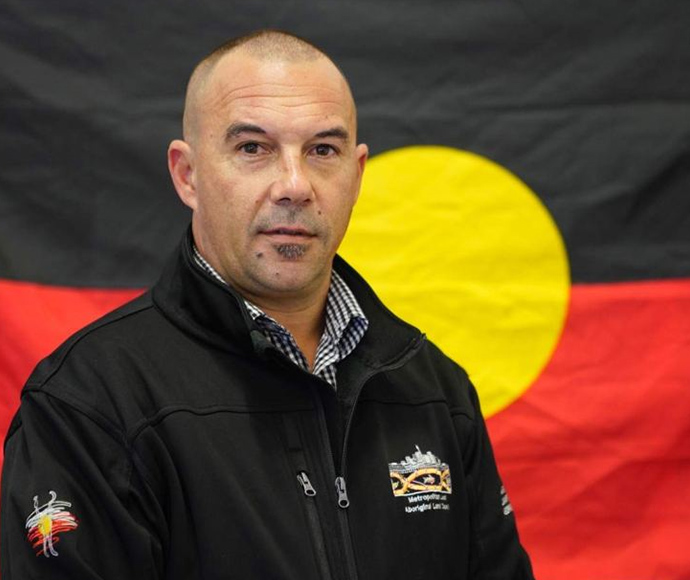 Nathan Moran is featured in the centre of the photo wearing a shirt and jacket, with the Aboriginal flag proudly sitting as the backdrop of the image.