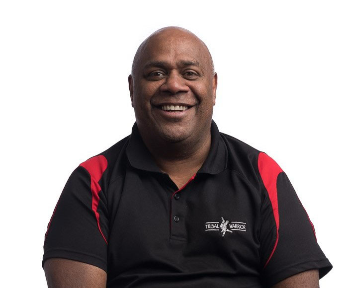 Shane Phillips is wearing a black and red polo with the words 'tribal warrior' on the chest. He is smiling at the camera with a plain white background.
