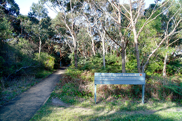 Entrance to the Jennifer Street Broadwalk at Kamay-Botany National Park in La Perouse, surrounded by trees and lush greenery.