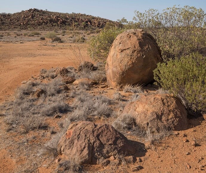 A photo of Dead Horse Gully Campground in Sturt National Park, located in the arid north-west corner of New South Wales. The scene features three rocks on rocky soil of the outback land, next to some vegetation, under a clear sunny sky.