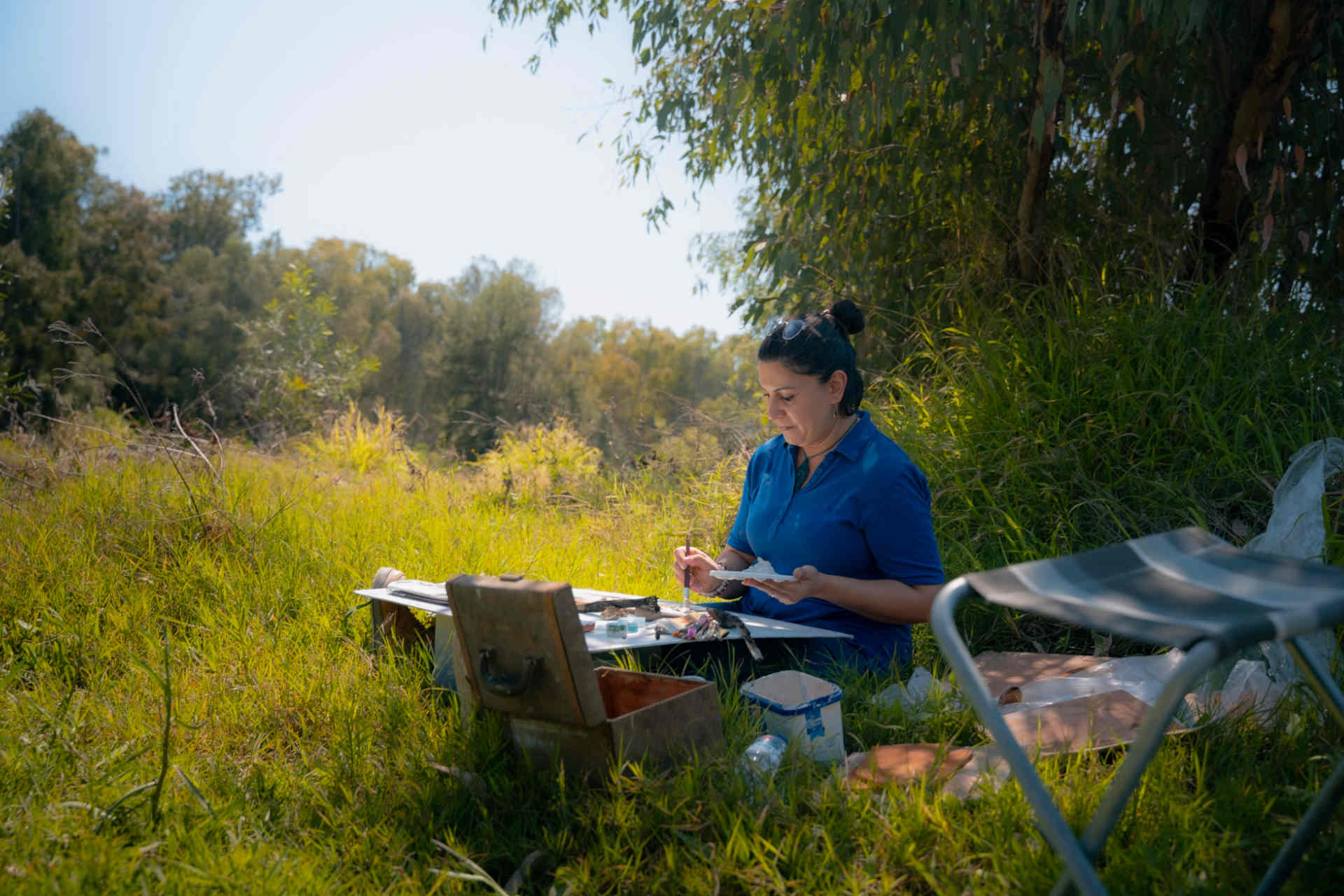 A person sitting cross-legged in the grass with a small art-making set-up around them, holding a palette and paint brush, among the riverside vegetation