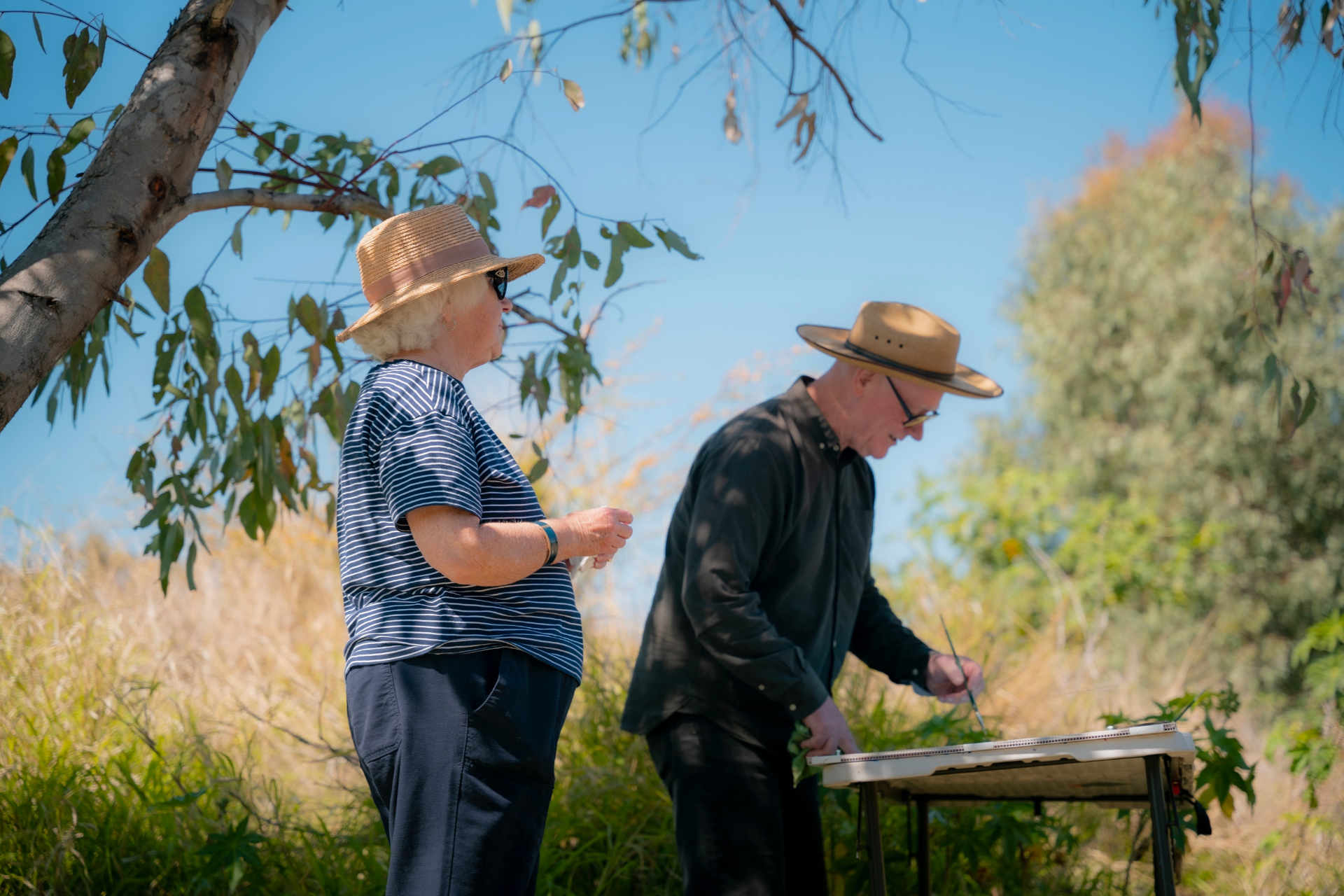 Two elderly people wearing hats and glasses standing under a gum tree, one of whom is painting onto a canvas