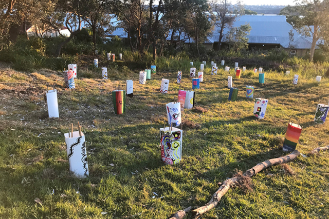 A grassy green patch of land with many cylinders standing upright with stakes surrounding new plantings. The cylinders have Aboriginal artwork on them  and extend across the land into towards some trees and blue water.